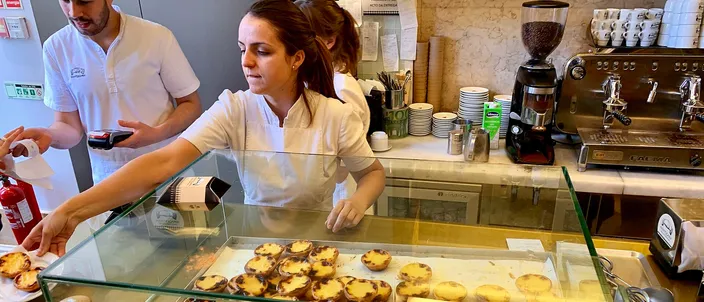 The counter of a Lisbon pastelaria: a glass case filled with rows of scorch-topped pasteis de nata, white-shirted staff plating tarts behind it, an espresso machine and stacked cups against the wall.