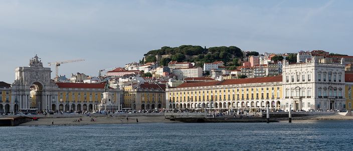 Praça do Comércio seen from the Tagus: the yellow arcaded riverfront square with its triumphal arch and equestrian statue, the green castle hill and terracotta rooftops rising behind, water in the foreground.