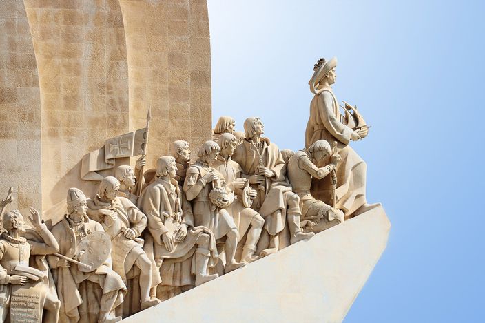 A close view of the carved stone prow of the Padrao dos Descobrimentos against a clear blue sky: a robed figure holding a small sailing ship stands alone at the point, with a file of carved figures in period dress ranged along the sloping edge behind him.
