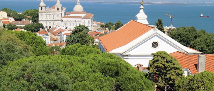 Seen from a height over pine tops: a white church with twin bell towers and a dome on the left, a second white church with an orange-tiled roof and a rose window on the right, tightly packed terracotta rooftops between them, and the wide blue Tagus estuary behind under a clear sky.