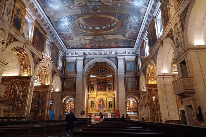 The interior of a large church looking towards the altar: a flat painted ceiling above with an illusionistic dome and coffering, gilded chapels down both side walls, a tall gilt-and-painted high altarpiece at the far end, dark wooden pews below, and a few small figures standing in the nave.
