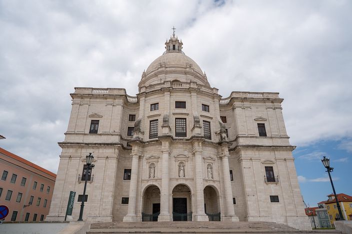 The white stone west front of a domed baroque church photographed from below: a tall central bay with paired columns and two arched doorways, curved wings either side, statues in niches, and a ribbed dome on a drum rising behind, against a broken cloudy sky.