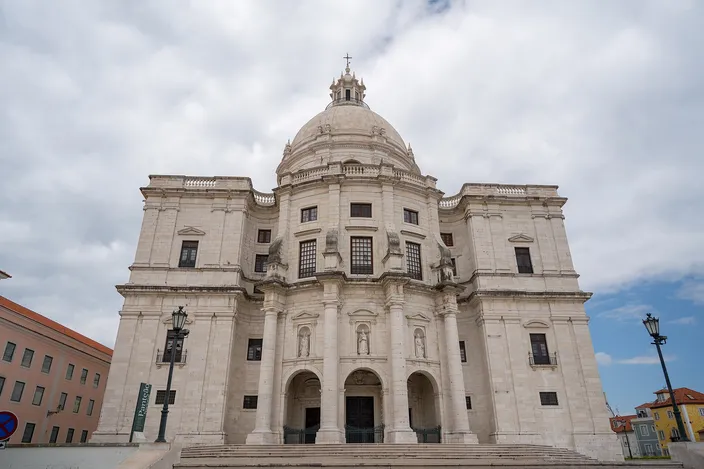 The white stone west front of a domed baroque church photographed from below: a tall central bay with paired columns and two arched doorways, curved wings either side, statues in niches, and a ribbed dome on a drum rising behind, against a broken cloudy sky.