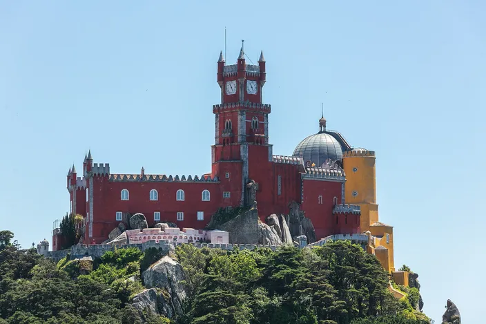 A palace of red and yellow blocks with battlements, a red clock tower and a gray stone dome standing on a bare rock outcrop above dense green woodland, under a clear pale blue sky.