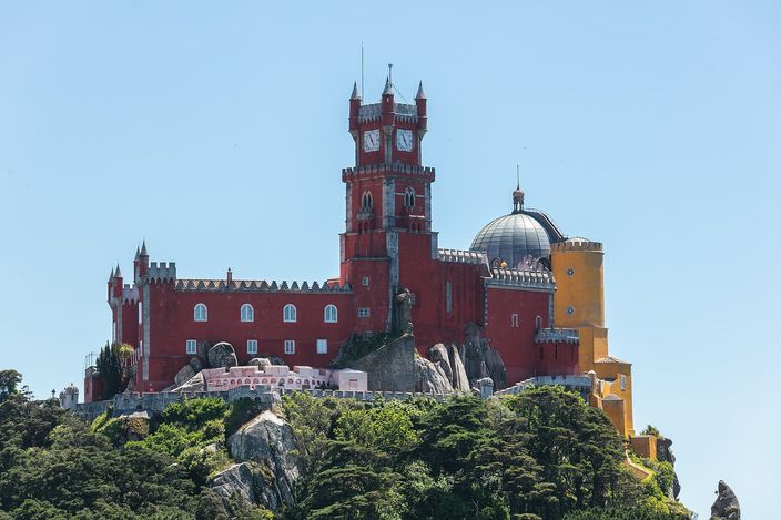 A palace of red and yellow blocks with battlements, a red clock tower and a grey stone dome standing on a bare rock outcrop above dense green woodland, under a clear pale blue sky.