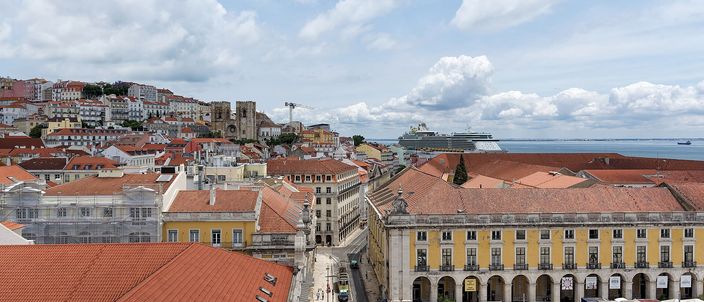 Looking east from a high archway over Lisbon at midday: a hillside of tightly packed terracotta roofs on the left carries the twin square towers of the cathedral, a straight pedestrian street runs down the centre, a long yellow arcaded wing with white cafe awnings closes the right, and a white cruise ship sits on the pale estuary under a cloudy sky.