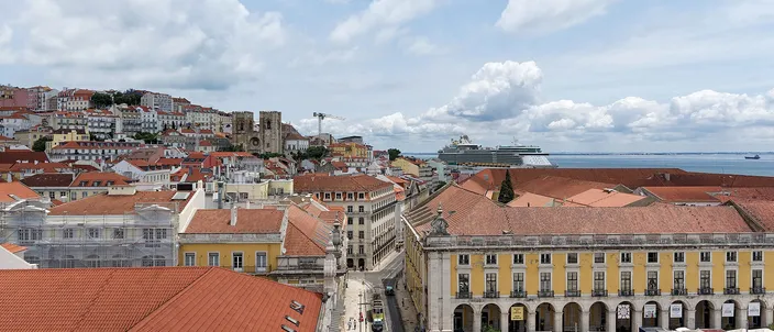Looking east from a high archway over Lisbon at midday: a hillside of tightly packed terracotta roofs on the left carries the twin square towers of the cathedral, a straight pedestrian street runs down the center, a long yellow arcaded wing with white cafe awnings closes the right, and a white cruise ship sits on the pale estuary under a cloudy sky.