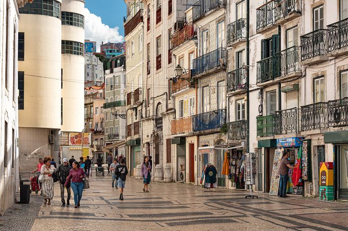 A wide cobbled street in Mouraria climbing into the hills, lined with tiled tenement facades and wrought-iron balconies, people walking on the polished calçada past small shops under a blue sky