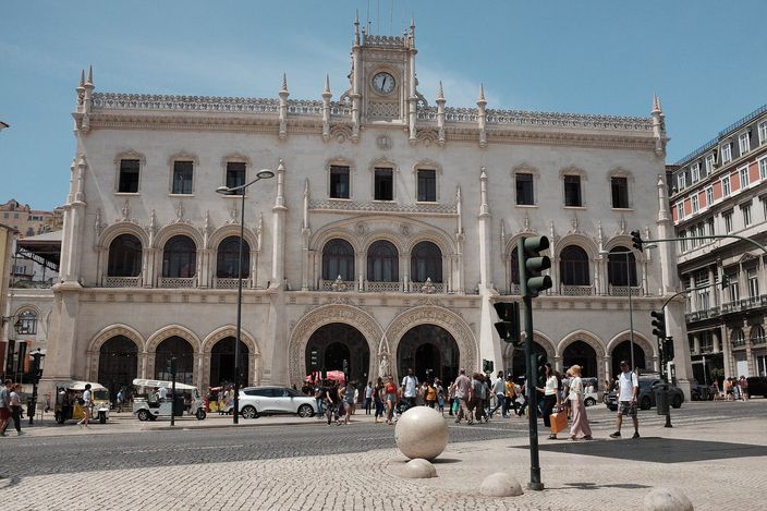 The ornate Manueline facade of Rossio railway station in Lisbon, with its twin horseshoe-arch doorways and clock, fronted by a busy pedestrian crossing and a stone bollard in the foreground under bright sun