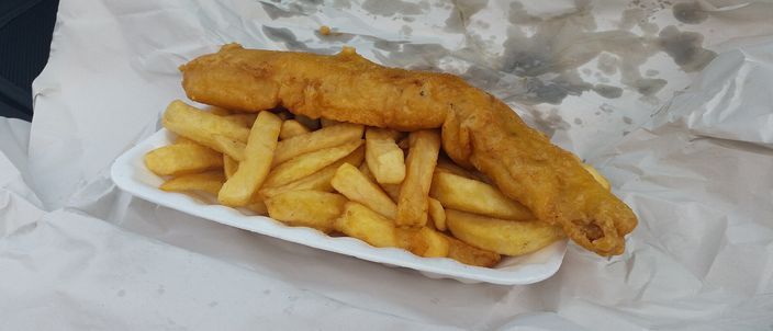 A battered fried white fish fillet with a heap of thick-cut chips, served on greaseproof paper in an open paper wrapping.