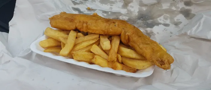 A battered fried white fish fillet with a heap of thick-cut chips, served on greaseproof paper in an open paper wrapping.