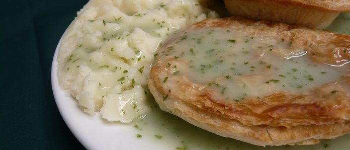 A white plate holding two golden pastry pies alongside a scoop of mashed potato, covered in pale green parsley liquor.