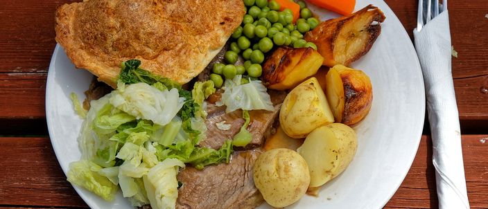 A plate of roast beef with a large Yorkshire pudding, roast potatoes, peas, carrot and greens, on a wooden pub table.