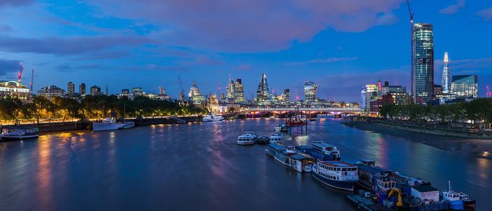 The Thames at blue hour seen from Waterloo Bridge, moored barges and pleasure boats in the foreground, Blackfriars Bridge crossing mid-frame and the City of London skyline lit on the north bank