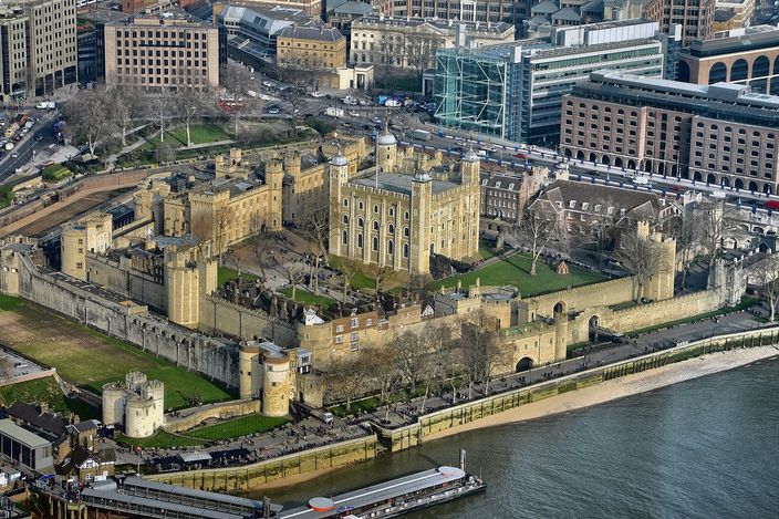 Sunlit aerial view of the whole Tower of London enclosure from above: the square White Tower with its four turrets in the centre, two concentric rings of battlemented curtain wall around it with smaller drum towers, the grassed dry moat outside them, the riverside wharf and the brown Thames along the bottom, and modern City offices pressed up against it at the top