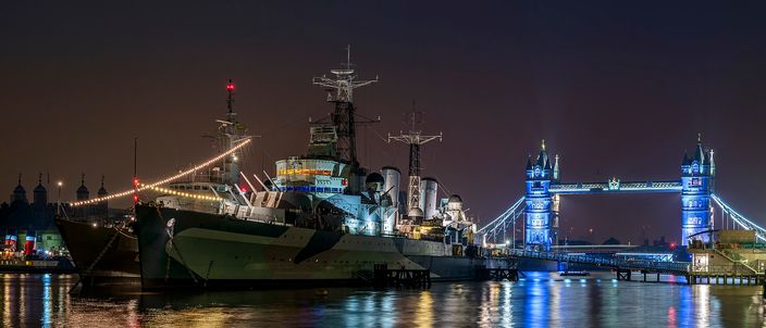 The grey warship HMS Belfast moored on the Thames at night, floodlit and strung with deck lights, its gun turrets trained forward; Tower Bridge lit blue behind it on the right, and the pale turrets and flagpoles of the Tower of London on the far bank at the left, all reflected in the black water