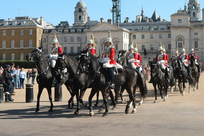 Mounted troopers of the Household Cavalry in red tunics and plumed helmets riding in line across the gravel of Horse Guards Parade, onlookers standing along the rail at the left and the pale stone and brick buildings of Whitehall behind them