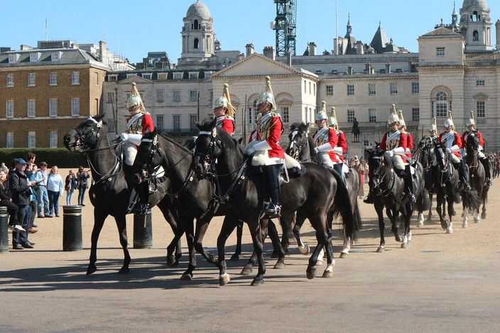 Mounted troopers of the Household Cavalry in red tunics and plumed helmets riding in line across the gravel of Horse Guards Parade, onlookers standing along the rail at the left and the pale stone and brick buildings of Whitehall behind them