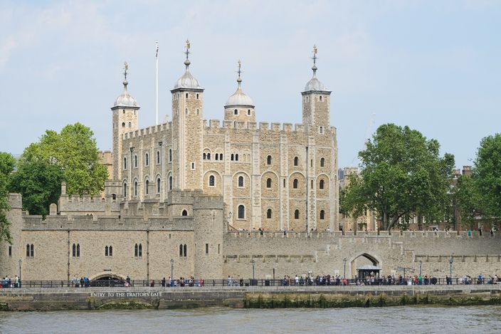 The Tower of London seen from the Thames on a clear day, the White Tower with its four corner turrets rising behind the crenellated outer wall, trees along the wharf and a line of visitors at the gate on the river front