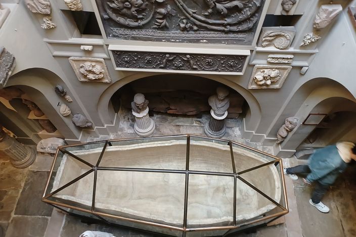 Looking down from a gallery into the basement of Sir John Soane's Museum, the alabaster sarcophagus of Seti I under a glazed case in the middle of the stone floor, the walls around it packed with carved fragments, capitals and relief panels, and a visitor on the stairs at the right