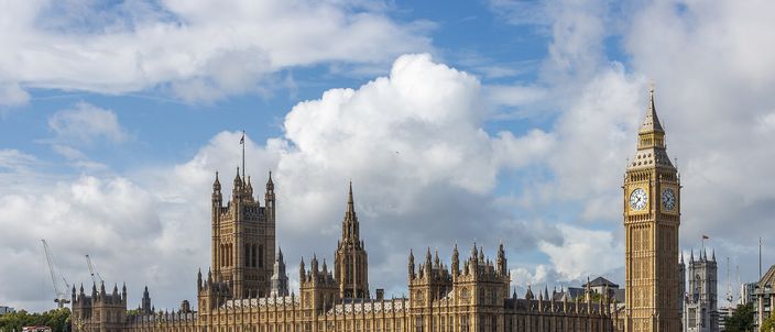 The Palace of Westminster stretched along the far bank of the Thames under bright piled cloud, the Elizabeth Tower and its clock face at the right-hand end, Westminster Bridge crossing the brown river in front of it with traffic on the deck