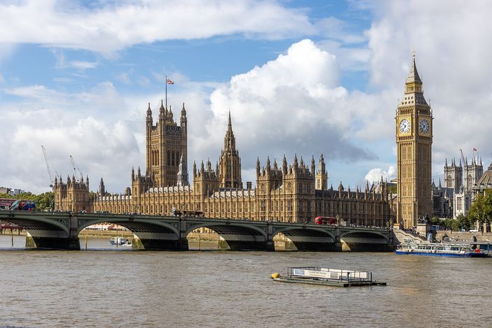 The Palace of Westminster seen across the Thames under cumulus cloud: the long Gothic river front, the Victoria Tower at the far end and the Elizabeth Tower carrying Big Ben's clock faces at the near one, red double-decker buses crossing Westminster Bridge in front and a barge on the brown water