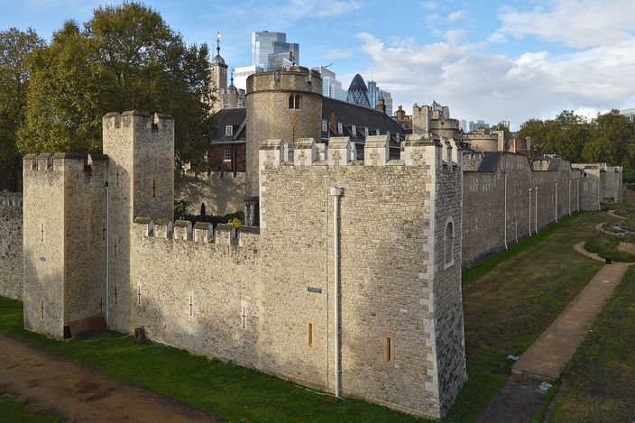 The outer curtain wall of the Tower of London in autumn: pale medieval stone, a round drum tower and battlemented walls above the grassed dry moat, with the glass towers of the City of London, including the Gherkin, rising directly behind