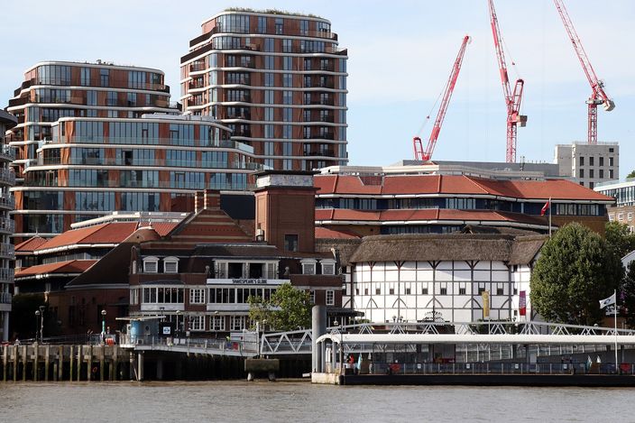 Shakespeare's Globe seen from the Thames: the lime-washed timber-framed drum with its conical thatched roof and the theatre's riverside frontage and pier, with modern apartment blocks and two red construction cranes on the skyline behind