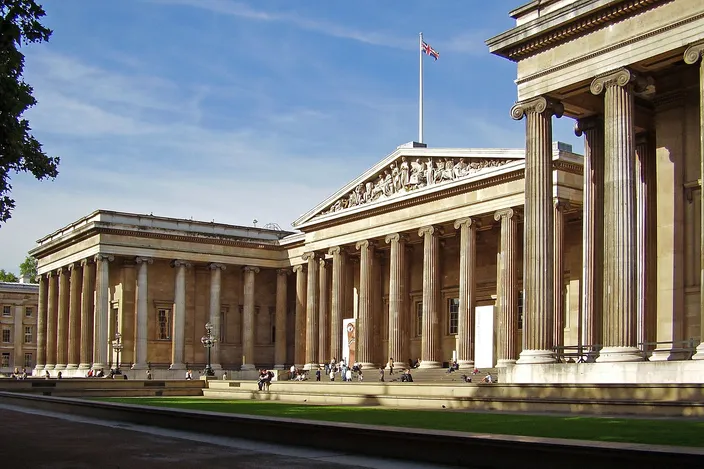 The main front of the British Museum from the north-east on a sunny day: a long Greek Revival colonnade of Ionic columns with a sculpted pediment over the central portico, the Union flag above it, and visitors on the forecourt steps