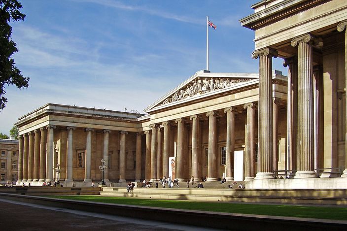 The main front of the British Museum from the north-east on a sunny day: a long Greek Revival colonnade of Ionic columns with a sculpted pediment over the central portico, the Union flag above it, and visitors on the forecourt steps