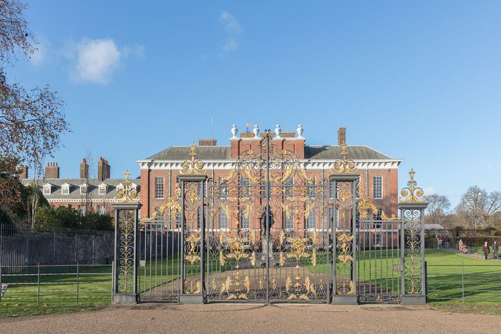 The south front of Kensington Palace under a clear blue sky seen through open gilded ironwork gates: a two-storey red-brick Baroque range with stone dressings and white urns along the parapet, lawn and gravel in front