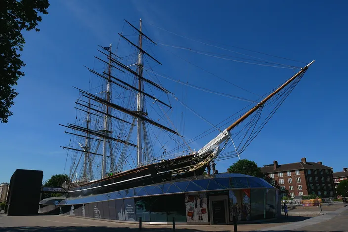 The Cutty Sark in its Greenwich dry dock against a deep blue sky: the clipper's dark hull raised above a glazed canopy at ground level, three masts with full standing rigging, and the bowsprit reaching out to the right over the sidewalk