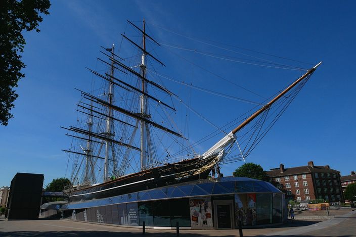 The Cutty Sark in its Greenwich dry dock against a deep blue sky: the clipper's dark hull raised above a glazed canopy at ground level, three masts with full standing rigging, and the bowsprit reaching out to the right over the pavement