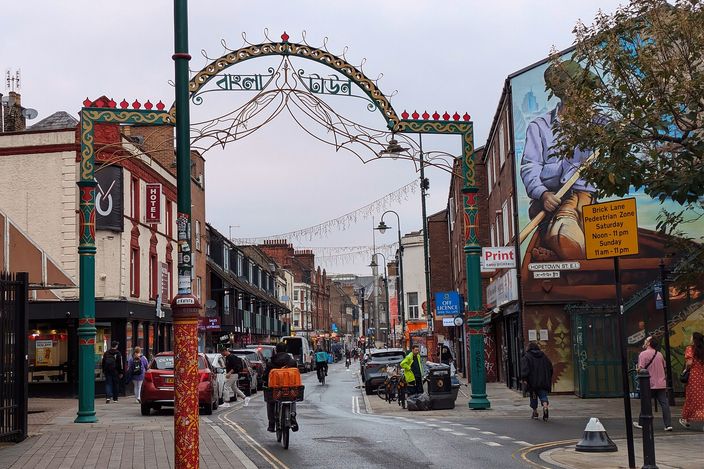 Brick Lane looking south on a damp day: a decorative steel arch spanning the street carrying the name in Bengali script, lamp posts painted in green, red and gold, shopfronts and a large wall mural along both sides, a cyclist and traffic on the wet road