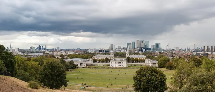 The view from the hill below the Royal Observatory in Greenwich: the lawns of Greenwich Park in the foreground, the white classical ranges of the Old Royal Naval College with the Queen's House in the gap between them below, and the towers of Canary Wharf and the City of London on the skyline under heavy cloud