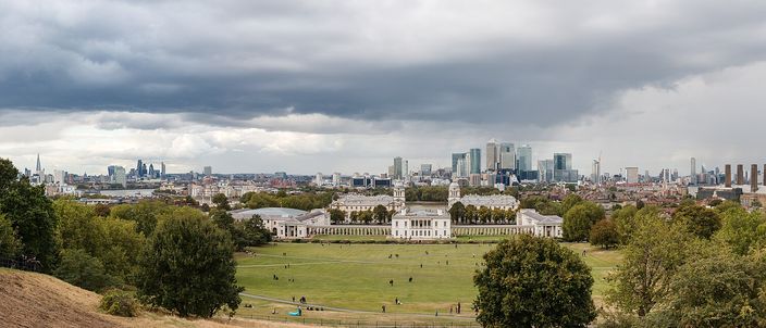 The view from the hill below the Royal Observatory in Greenwich: the lawns of Greenwich Park in the foreground, the white classical ranges of the Old Royal Naval College with the Queen's House in the gap between them below, and the towers of Canary Wharf and the City of London on the skyline under heavy cloud