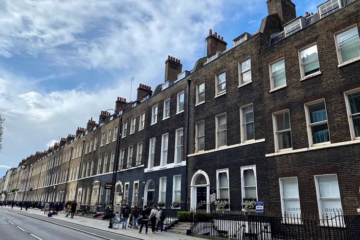 A long row of four-storey brown-brick Georgian terraced houses along Bloomsbury Street, white-painted sash windows and doorways, black iron railings and chimney stacks against a cloudy sky, with people walking on the pavement