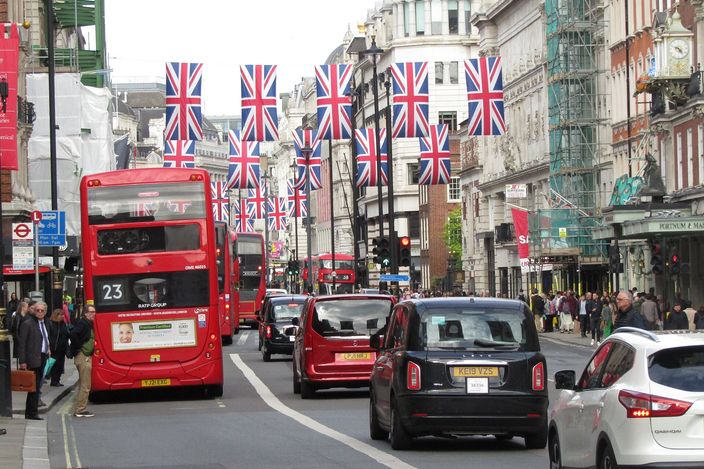 A red double-decker bus in the bus lane and black London taxis on Piccadilly, beneath rows of Union Jack flags strung across the street with shopfronts and scaffolding along the sides