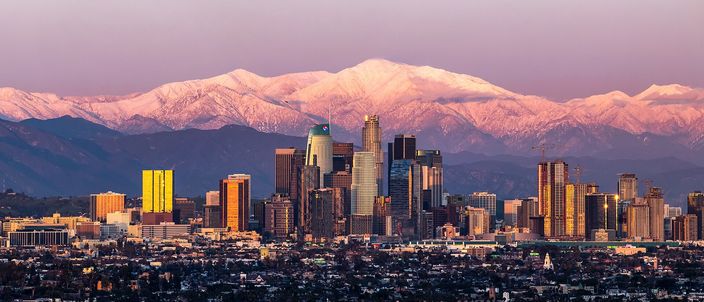 The Downtown Los Angeles skyline at sunset glowing pink, backed by the snow-capped San Gabriel Mountains with Mount Baldy rising above the towers