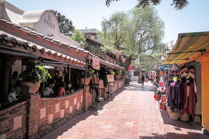 A brick-paved lane in Los Angeles under awnings and a pepper tree, stalls hung with embroidered dresses on the right and a tiled arcade with people eating at tables on the left