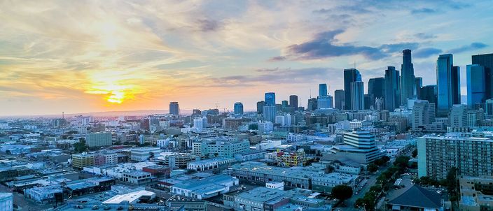 Downtown Los Angeles from the air at sunset: the tower cluster standing on the right against a warm sky, the low city running flat to a setting sun on the left horizon