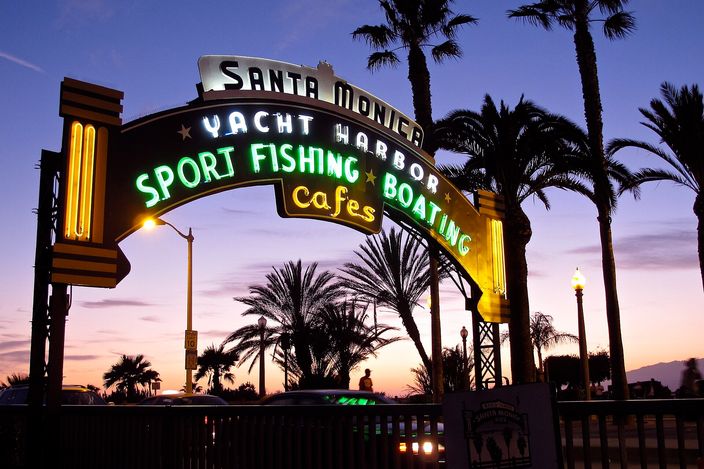 The Santa Monica Pier's neon gateway arch lit at dusk, reading 'Santa Monica Yacht Harbor, Sport Fishing, Boating, Cafes', with palm trees and a violet and orange sky behind it