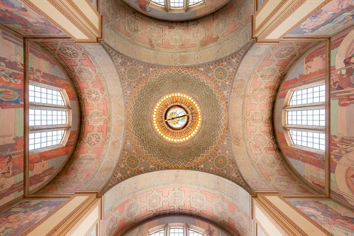 Looking straight up into the rotunda of the Los Angeles Central Library: a chandelier ringed with zodiac signs hanging from a mosaic dome, painted murals filling the four vaults around it and arched windows on each side