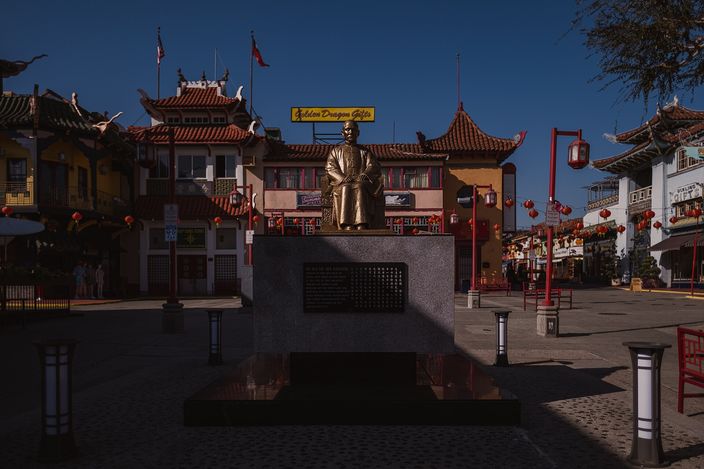 Los Angeles Chinatown's Central Plaza in low sun: pagoda-roofed buildings, a bronze statue on a plinth, red lanterns strung overhead and a neon 'Golden Dragon Gifts' sign