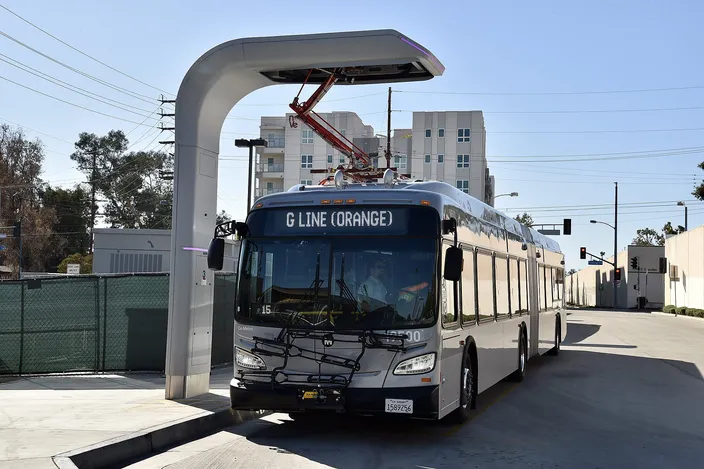 A silver New Flyer articulated LA Metro bus with 'G LINE (ORANGE)' on its destination sign, parked under an overhead charging arm at North Hollywood station on a clear day, apartment blocks behind