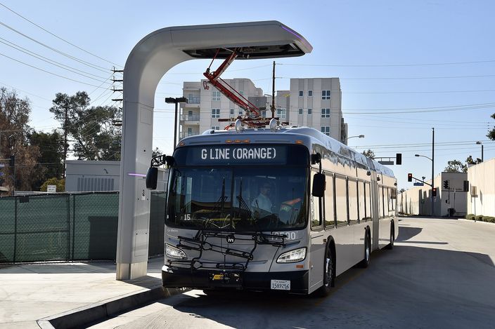 A silver New Flyer articulated LA Metro bus with 'G LINE (ORANGE)' on its destination sign, parked under an overhead charging arm at North Hollywood station on a clear day, apartment blocks behind