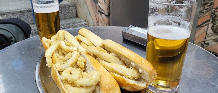 Two crusty bread rolls filled with rings of fried squid on a metal tray at a bar counter, flanked by two glasses of draught beer