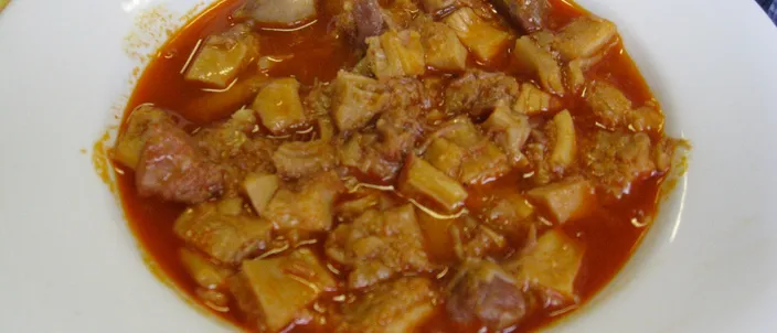 A white plate of callos: chunks of tripe and meat in a thick reddish-orange paprika sauce, with bread at the edge of the frame