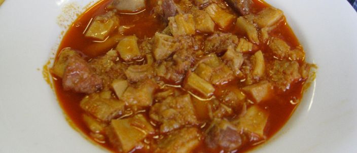 A white plate of callos: chunks of tripe and meat in a thick reddish-orange paprika sauce, with bread at the edge of the frame