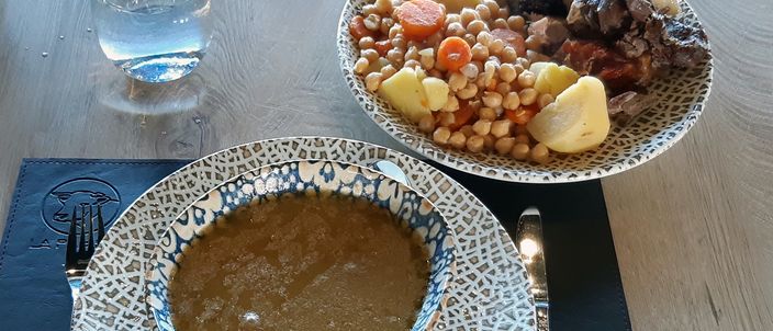 A bowl of golden cocido broth in the foreground and, behind it, a plate of chickpeas with potato, carrot and several boiled meats